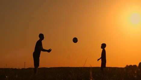 Vater-Und-Sohn-Spielen-Fußball-Im-Park-Bei-Sonnenuntergang,-Silhouetten-Vor-Dem-Hintergrund-Einer-Zeitlupenaufnahme-Einer-Hellen-Sonne.